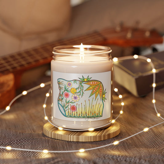 A lit candle in a clear glass jar sits on a round wooden coaster, surrounded by warm white fairy lights. The jar has a white label with a colorful illustration of a bird and flowers.
