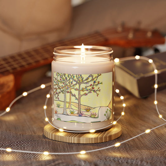A lit candle in a clear glass jar sits on a round wooden coaster, wrapped with warm white fairy lights. The jar features a label with a whimsical illustration of two trees in a snowy landscape.