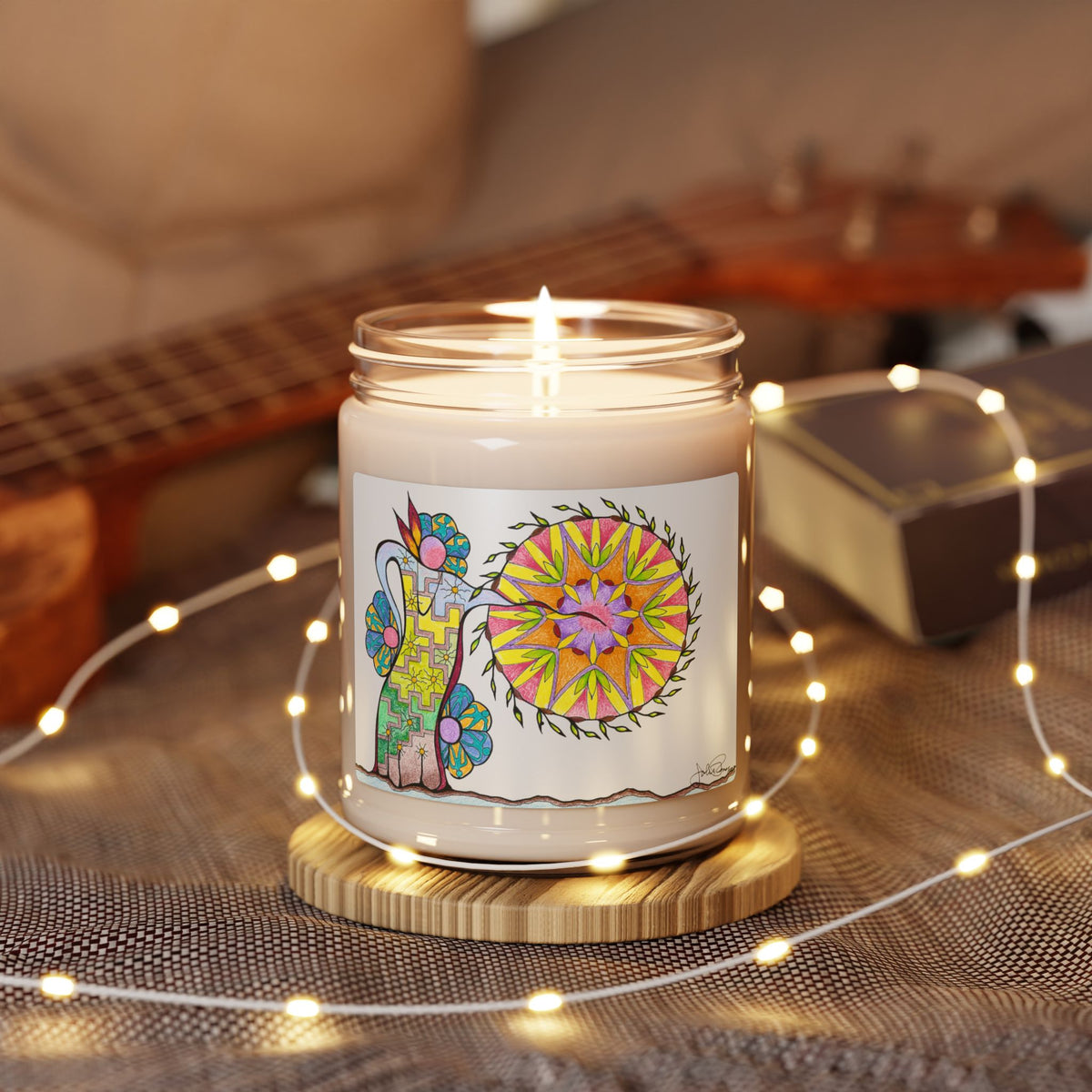 A lit candle in a clear glass jar sits on a round wooden coaster, surrounded by warm fairy lights. The jar has a white label featuring a colorful illustration of an interpretation of a Native American Pow Wow.